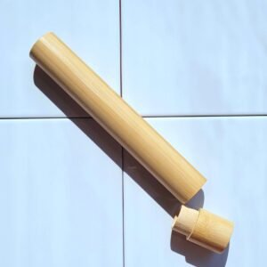 Un étui à brosse à dents en bambou, lisse et de couleur claire, est posé sur un fond de carrelage bleu pâle. Le corps principal est allongé et cylindrique, et son capuchon est partiellement détaché, révélant l'intérieur creux. L'étui est éclairé par une lumière vive qui projette une ombre sur le carrelage.