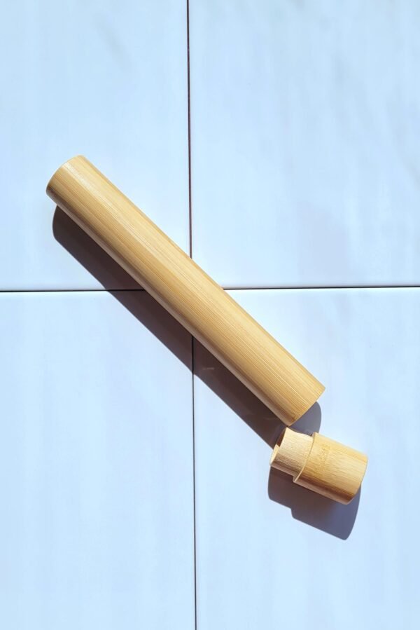 Un étui à brosse à dents en bambou, lisse et de couleur claire, est posé sur un fond de carrelage bleu pâle. Le corps principal est allongé et cylindrique, et son capuchon est partiellement détaché, révélant l'intérieur creux. L'étui est éclairé par une lumière vive qui projette une ombre sur le carrelage.