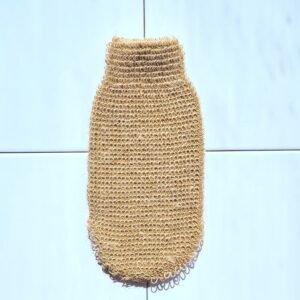 Un gant de gommage en fibres de jute tressées, de couleur beige-marron, est posé à plat sur un carrelage blanc avec des lignes de joints grises. Le gant a une forme ovale, plus large en bas et se rétrécissant vers une ouverture en haut. Sa texture est visible, composée de petites boucles de fil.